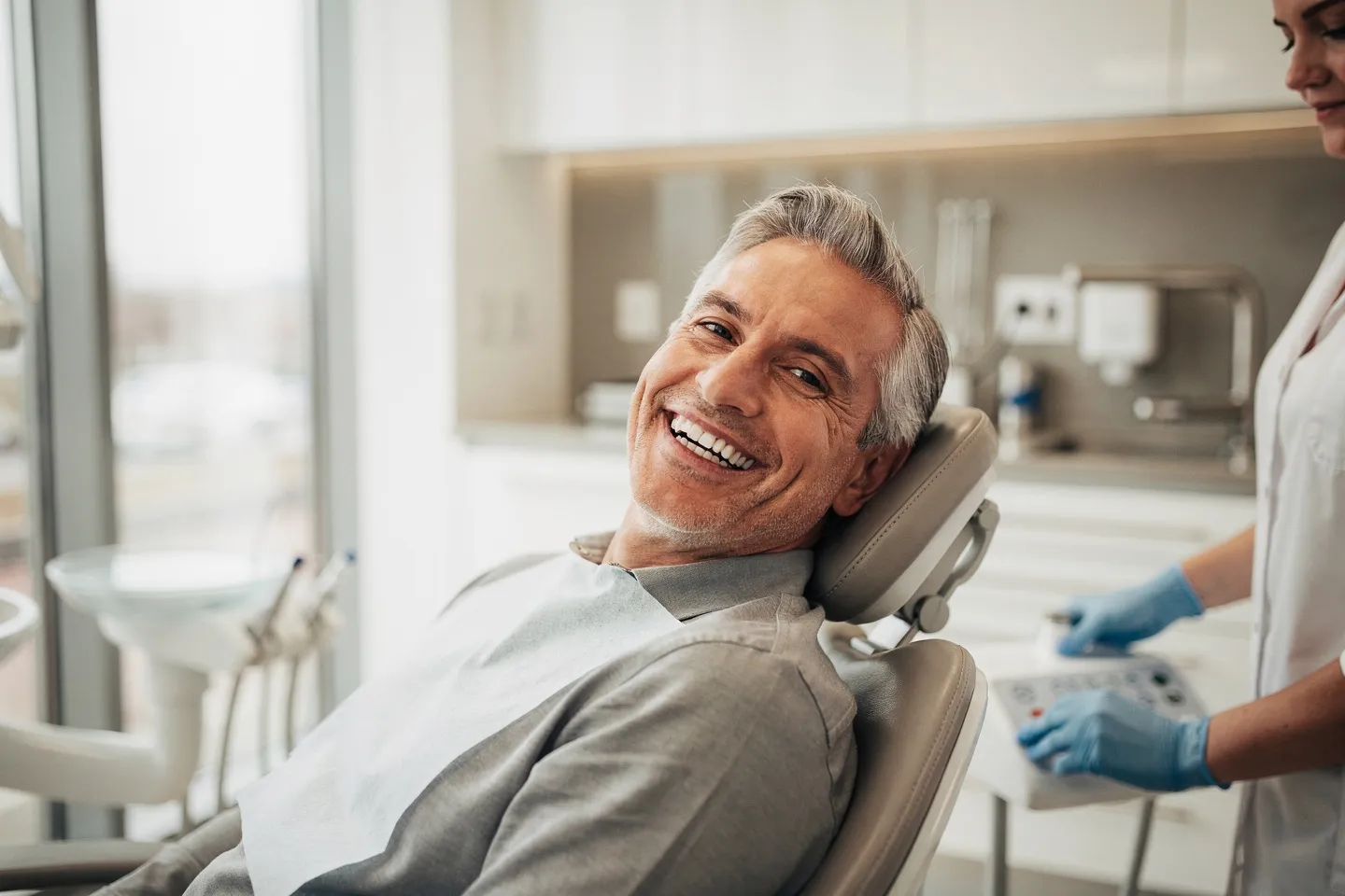 Ein lächelnder Patient mittleren Alters sitzt auf einem modernen Zahnarztstuhl und zeigt perfekte Zähne