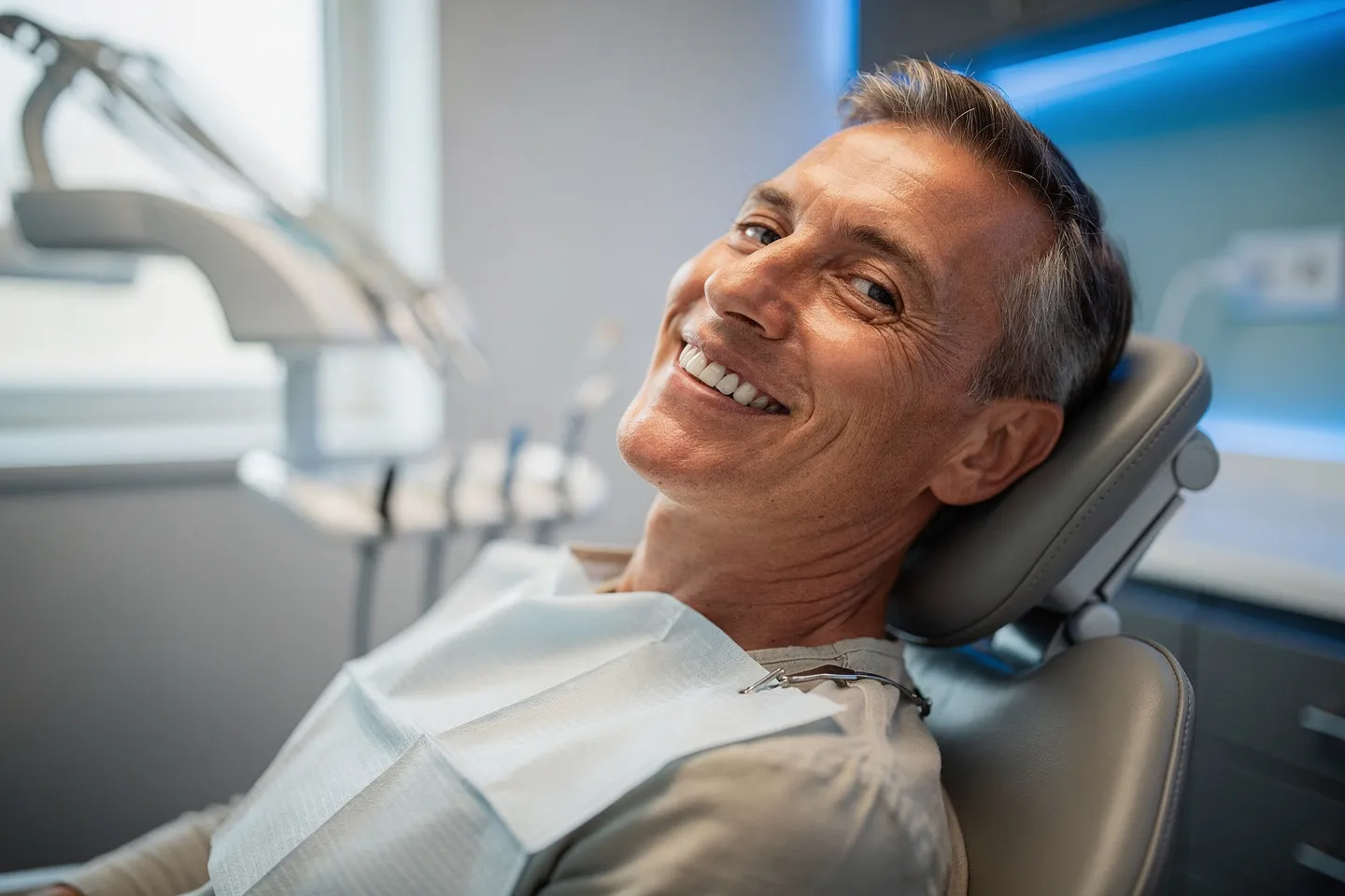 Ein lächelnder Patient mittleren Alters sitzt auf einem modernen Zahnarztstuhl mit perfekten weißen Zähnen