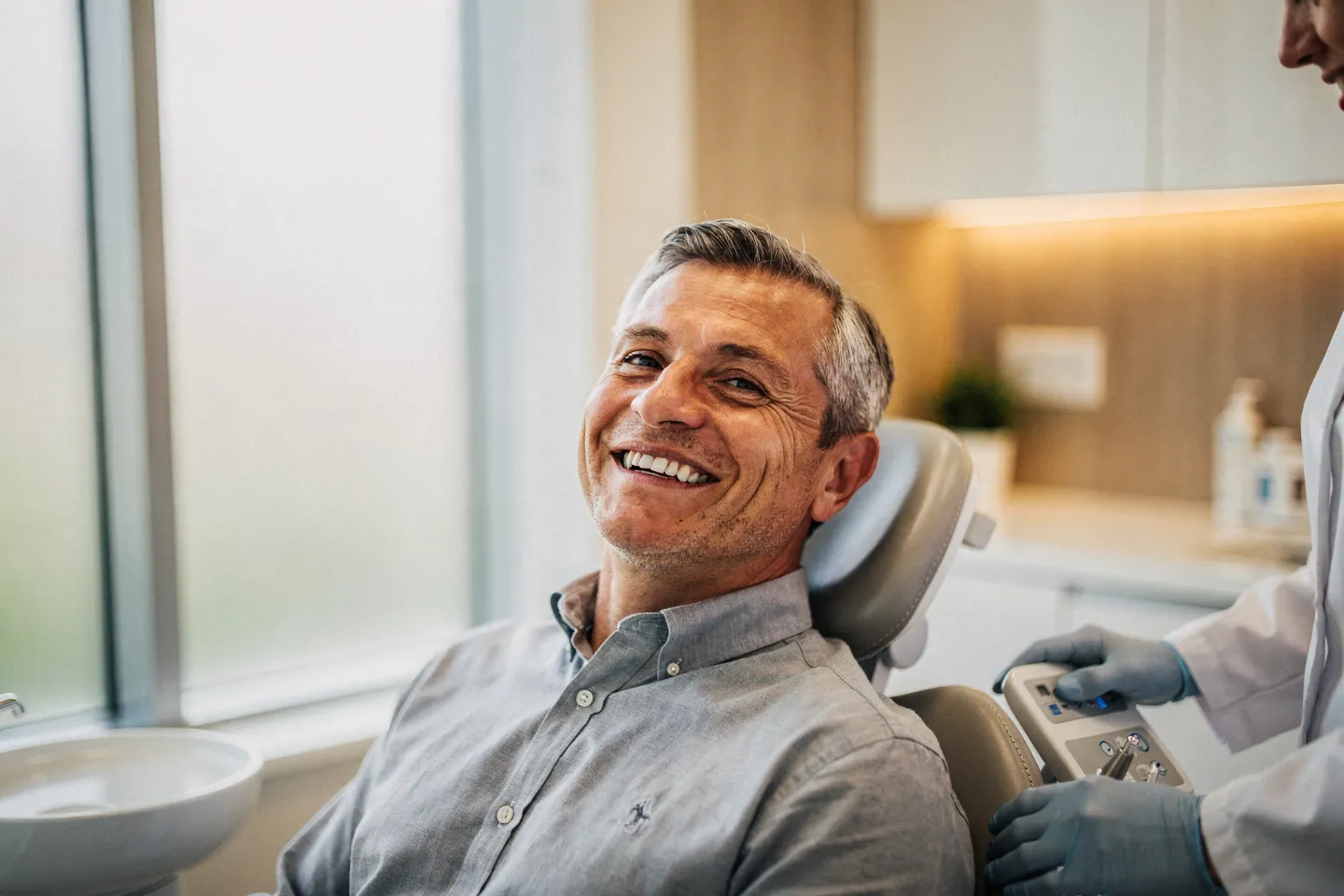 Ein lächelnder Patient mittleren Alters sitzt entspannt auf einem modernen Zahnarztstuhl in einer hellen Praxis