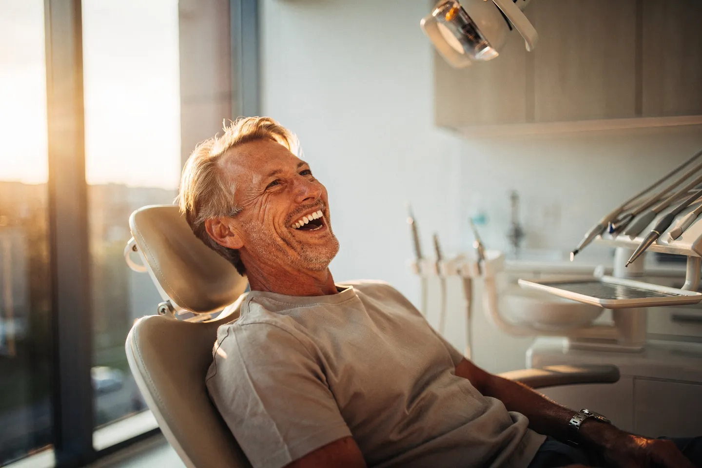 Ein lachender Patient mit weißen Zähnen sitzt in einem modernen Zahnarztstuhl bei Sonnenlicht